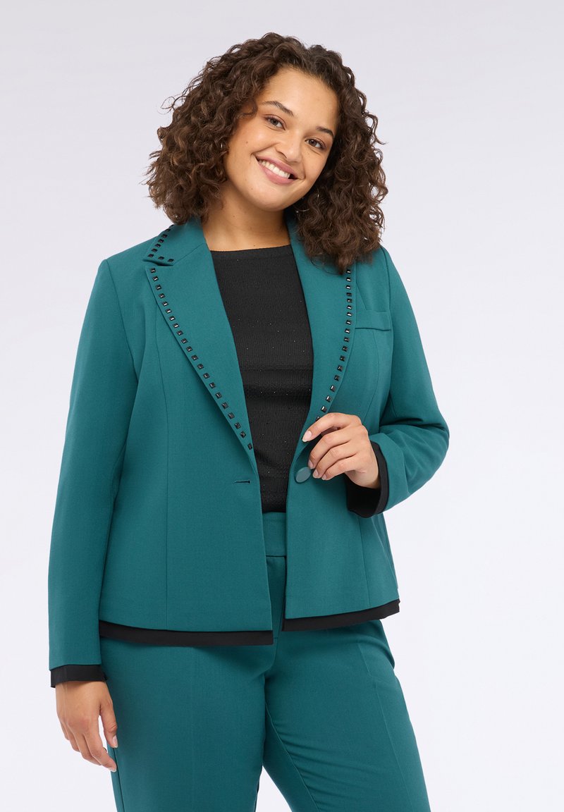 Blazer teal con accenti neri e dettagli con borchie lungo il colletto, abbinato a un top nero e pantaloni coordinati, caratterizzati da una vestibilità strutturata.