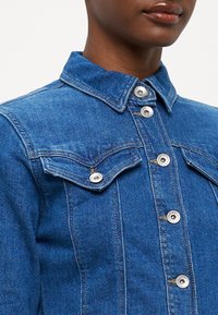 Denim jakna z značilnim ovratnikom, srebrnimi gumbi na pritisnik in žepi na prsih. Tkanina je v srednje modri barvi z rahlo variacijo teksture.