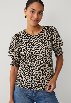 Kvinde iført en kortærmet leopardprintbluse med puffede ærmer og blå jeans, smilende let og holdende i blusens kant.