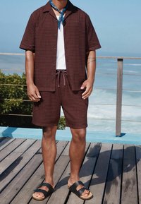 Chemise marron à manches courtes avec boutons et motif texturé, associée à un short assorti, et sandales noires avec semelle en liège.