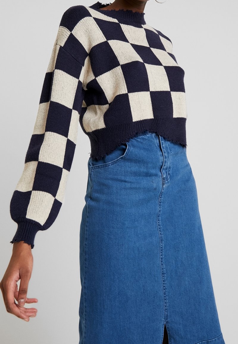 Pull en maille damier bleu marine et crème avec un design court et un ourlet effiloché, associé à une jupe en denim avec une fente à l'avant.
