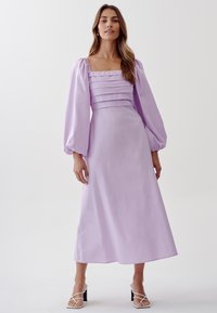 Lilac long dress with a square neckline, pleated bodice, puffed sleeves, and an A-line silhouette, paired with white strappy heels.