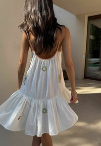 Biele šaty bez rukávov s viacvrstvovou sukňou, zdobené farebnou kvetinovou výšivkou a otvoreným chrbtom do tvaru výstrihu. Ľahká tkanina s prúdiacim dizajnom.