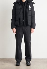 Veste matelassée noire avec capuche, arborant un logo sur la poitrine, associée à une chemise noire à boutons et un pantalon noir ample, complétée par des baskets noires.