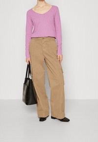 Pitkähihaisessa, V-kaula-aukkoisessa vaaleanpunaisessa paidassa on yhdistetty vaaleat cargo-housut, joissa on sivutaskut; malli pitää mustaa olkalaukkua ja hänellä on mustat kengät.