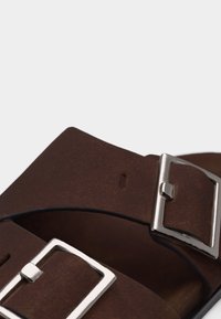 Deux brides de sandales en cuir marron qui se chevauchent, chacune avec une boucle rectangulaire en métal argenté, sur un fond clair.