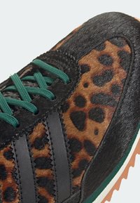 Chaussure de sport avec fourrure à imprimé léopard, bandes en cuir noir, lacets vert foncé et semelle blanche avec une semelle extérieure marron.