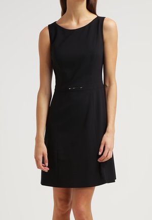 Woman wearing a sleeveless black knee-length dress with a round neckline and thin belt detail at the waist against a plain background.