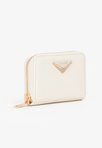 Portafoglio compatto color crema con zip dorata e placca logo dorata a forma triangolare sul davanti, fotografato su sfondo bianco.