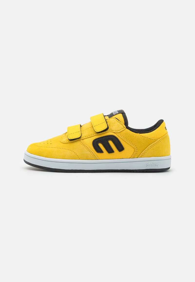Yellow suede sneaker with black accents, featuring two Velcro straps, rounded toe, and a grey rubber sole. Embossed branding on the side.