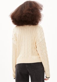 Pull en maille torsadée crème avec ourlet et poignets côtelés, présentant une coupe décontractée et un motif texturé. Porté avec un jean foncé.