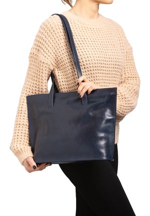 Woman wearing beige knitted sweater holding large navy blue leather tote bag with shoulder straps against white background.