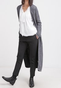 Woman wearing a long gray knitted cardigan, white blouse with bow, black slim pants, and black woven ankle boots standing with hands in pockets.