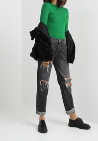 Grünes gestreiftes Langarmshirt, kombiniert mit ausgewaschenen schwarzen Jeans mit hoher Taille. Schwarze Kunstpelzjacke, über die Schultern gelegt, schwarze Schuhe.