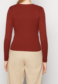 Ribbed top z długim rękawem w kolorze rdzawego pomarańczu, z okrągłym dekoltem i dopasowanym krojem. Połączony z jasnobeżowymi spodniami.
