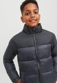 Črna puffer jakna z visokim ovratnikom, z zadrgo spredaj, kontrastnimi paneli in prešito teksturo. Oblikovana za toploto.