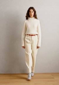 Pull torsadé crème à col roulé, pantalon blanc ample, ceinture rouge, bottines grises à motifs, debout sur un sol en bois devant un fond uni.