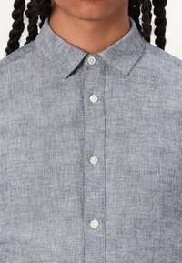 Lichtblauw linnen shirt met een kraag, voorzien van een knoopsluiting aan de voorkant en witte knopen. De stof heeft een gestructureerde, licht geweven uitstraling.