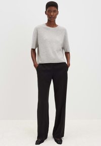 Femme se tenant debout les mains dans les poches, portant un pull gris à manches courtes, un pantalon noir à jams larges et des chaussures noires à bouts pointus.