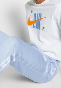 Bílé tričko s barevným potiskem "Nike AIR", oranžové logo Swoosh, zelená postava; světle modré kalhoty se stahovacími pásky a zipem po straně.