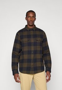 Korte mouwen geruite blouse met olijfgroene en marineblauwe vierkanten, gemaakt van zachte stof. Voorzien van een knoopsluiting aan de voorkant en twee borstzakken.