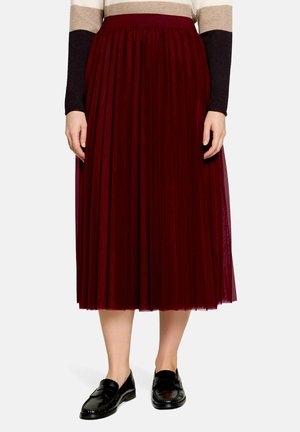 Kvinde iført et vinrødt plisseret midi-nederdel med sorte loafers og en langærmet sweater med beige, brune og sorte farveblokke.