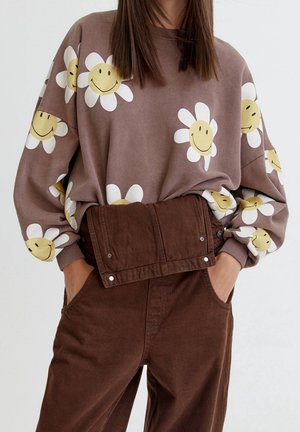 Braunes Sweatshirt mit großen weißen Gänseblümchen mit gelben Smileys, kombiniert mit hoch taillierter brauner Hose und Händen in den Taschen.