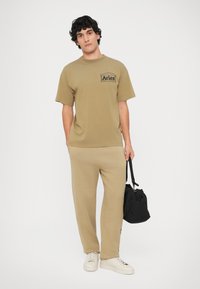 Bruine katoenen T-shirt met een logo op de linkerborst, gecombineerd met bijpassende joggingbroek en witte sneakers. Zwarte tas vastgehouden in de linkerhand.