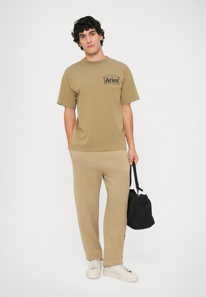 Bruine katoenen T-shirt met een logo op de linkerborst, gecombineerd met bijpassende joggingbroek en witte sneakers. Zwarte tas vastgehouden in de linkerhand.