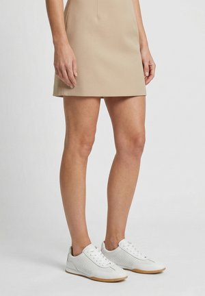 Žena v béžové minisukni a bílých perforovaných teniskách na šněrování stojí před jednobarevným pozadím.