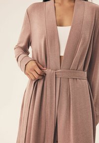 Jasnoróżowy kardigan o prążkowanej fakturze, z otwartym przodem i wiązanym w pasie. Długie rękawy i boczne drapowania podkreślają swobodny krój.