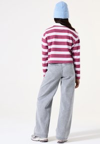 Gestreiftes Langarmshirt in Kastanienbraun, Rosa und Blau, kombiniert mit grauen, hochgeschnittenen, weiten Hosen. Strickmütze in Blau auf dem Kopf.