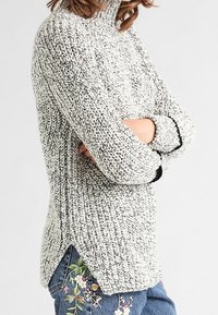 Chunky stickad tröja i grått och vitt med hög krage, sidofickor och svarta detaljer. Mjuk textur med avslappnad passform.