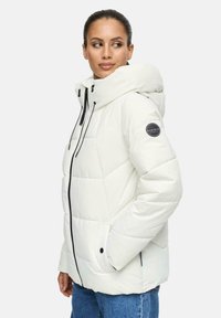 Vrouw in een witte gewatteerde jas met capuchon, zwarte ritssluiting en logo-opdruk op de mouw, met haar handen in de zakken, kijkend naar de zijkant.