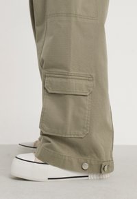Olivově zelené cargo kalhoty s postranním kapsou, rovný střih a látkové knoflíkové zapínání u kotníku, v kombinaci s bílými teniskami.