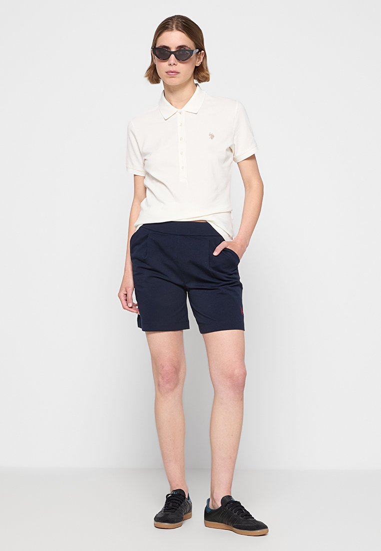 Polo shirt bianco con bottoni, abbinata a pantaloni corti blu navy con tasche. Sneakers scure e occhiali da sole neri completano l'outfit.