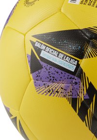 Geltonas futbolo kamuolys su tekstūruota paviršiumi, juodais ir violetiniais akcentais, bei užrašu "BALÓN OFICIAL DE LALIGA 2024/2025 sezonas."