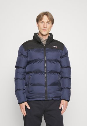 Uomo che indossa una giacca imbottita blu navy e nera con zip e tasche frontali, in piedi davanti a uno sfondo chiaro e uniforme.