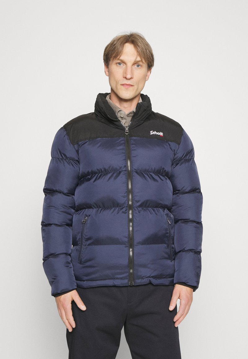 Homme portant une doudoune zippée bleu marine et noire avec des poches frontales, debout devant un fond clair uni.