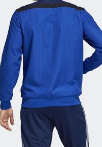 Blå atletisk jacka med långa ärmar, texturerad tyg, svarta detaljer på axlarna, elastisk nederkant och ärmslut. Enkelt design, inga mönster.