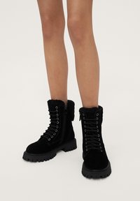 Botas de tobillo de ante negras con un collar de piel texturizada, cordones en la parte delantera, cremallera lateral y suela de goma gruesa con un patrón de relieve.