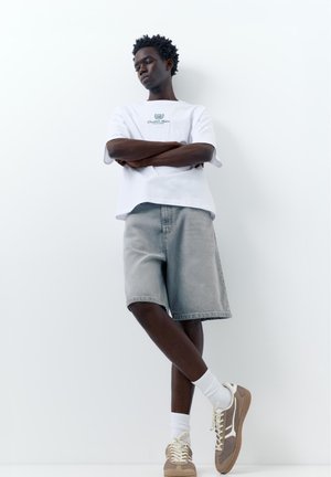 Tânăr cu brațele încrucișate purtând un tricou alb cu grafică, pantaloni scurți gri din denim, șosete albe și adidași bej, stând în fața unui perete alb.