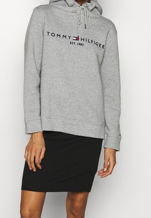 Kvinde iført en grå Tommy Hilfiger hættetrøje med logo og sort nederdel, stående foran en hvid baggrund.
