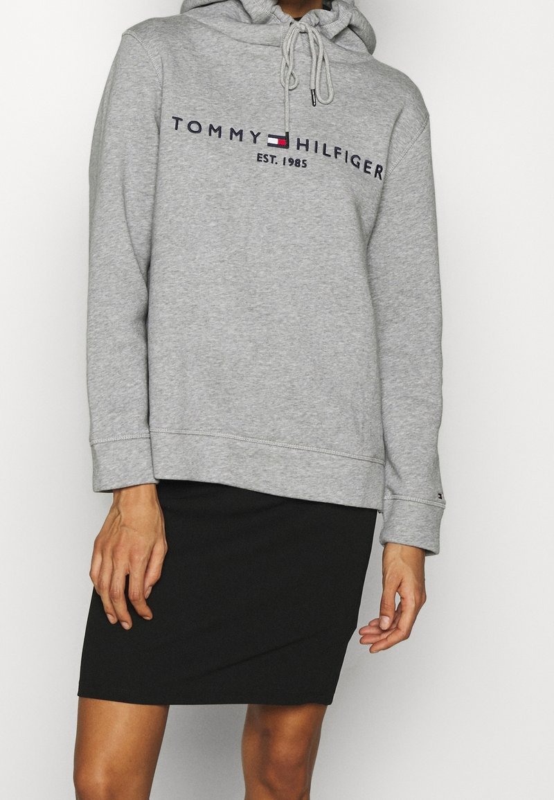 Femme portant un sweat à capuche gris Tommy Hilfiger avec logo et une jupe noire, debout devant un fond blanc uni.