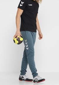 Sort kortærmet skjorte med hvidt logo; teal bukser med sølv chevron-detaljer; holder en gul og sort bold; iført sort og hvidt sneakers.