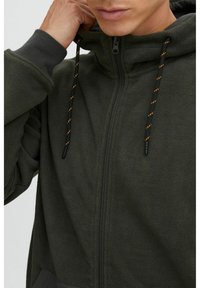 Chaqueta de forro polar de color verde oscuro con cremallera frontal, capucha ajustable y acentos en naranja contrastante en los cordones. Presenta una tela texturizada.
