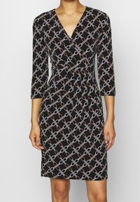 Robe portefeuille noire avec un motif géométrique de chaînes en blanc et cuivre, manches trois-quarts et taille ajustée. Texture du tissu lisse.