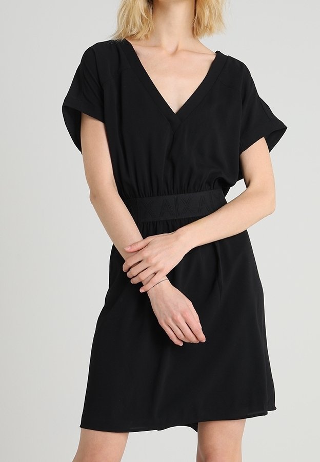 Femme portant une robe noire à manches courtes avec encolure en V et taille cintrée, debout les mains doucement jointes devant elle.