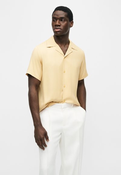 Homme portant une chemise à manches courtes jaune clair boutonnée et un pantalon blanc, debout avec une main dans la poche, regardant vers la gauche.