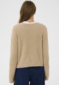 Smėlio spalvos pūkuotas megztinis ilgomis rankovėmis, apvalia iškirpte ir trumpa apačia. Audinys yra minkštas ir pūkuotas, šiek tiek laisvo kirpimo.